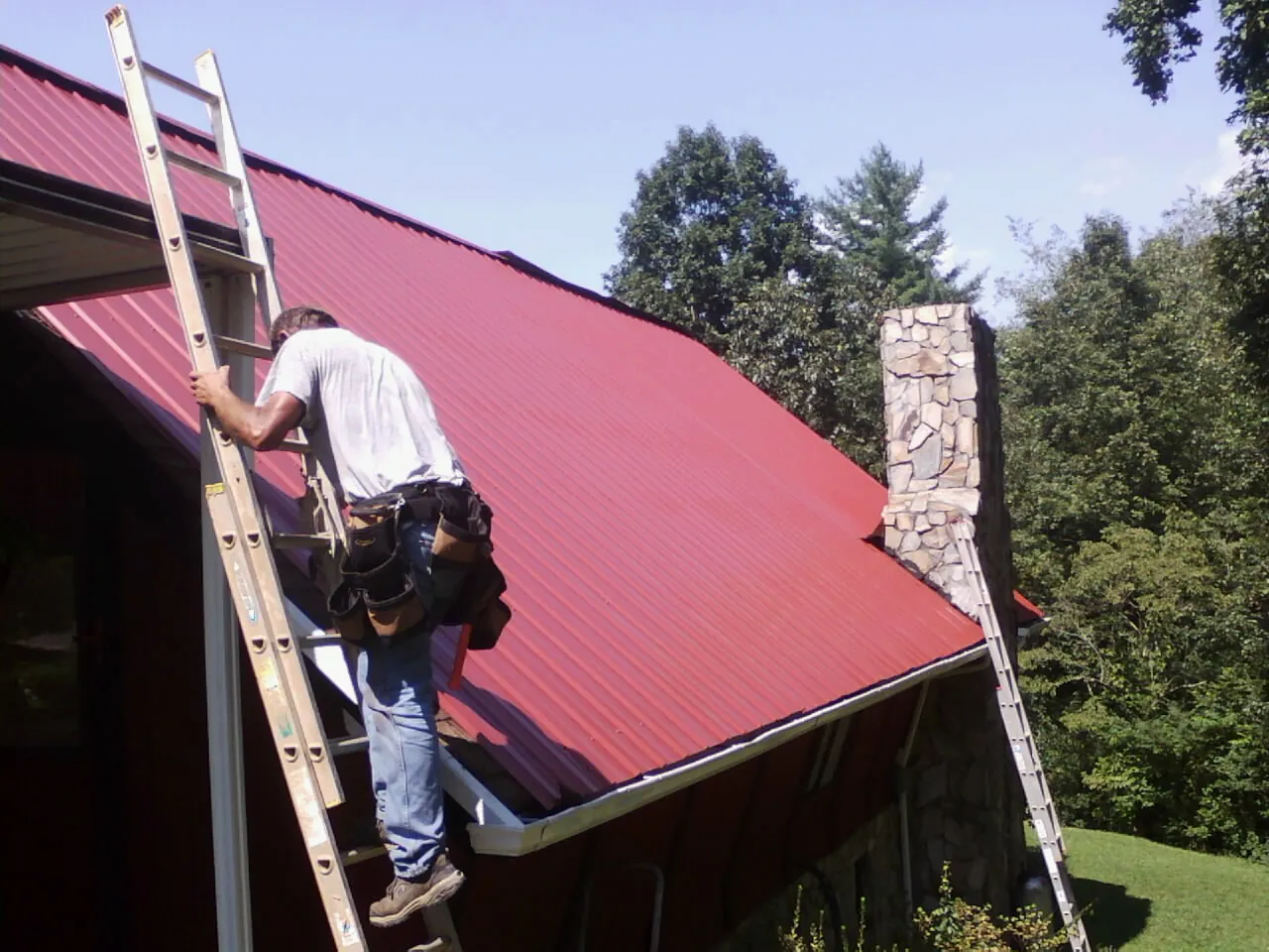 Roofer on a ladder beside a red metal roof and stone chimney — Soffit & Fascia Repair by Roofing Brothers in Burlingame